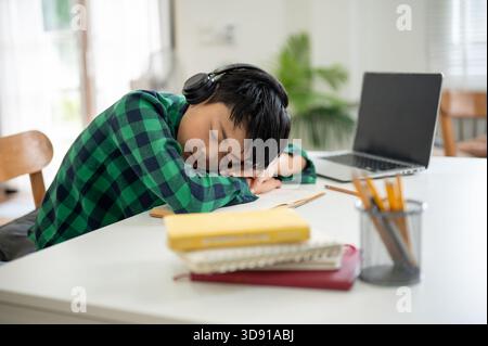 Asian kid elementary school boy wearing headphones sleeping over a book on table with laptop aside. Online Study, Doing homework, Internet surfing. Stock Photo