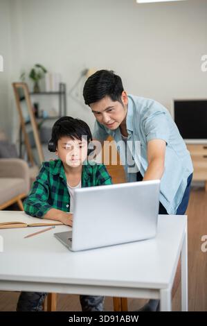 Asian father teaching kid elementary school boy wearing headphones while looking at laptop together. Online Study, Doing homework, Internet surfing. Stock Photo