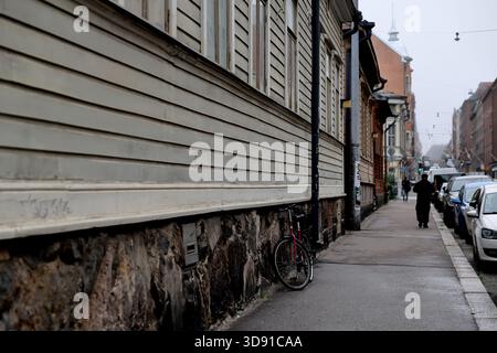 Old wooden residential building with bicycle leaning by stone foundation along city street with parked cars in Helsinki, Finland on a cloudy day. Stock Photo