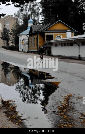 Male cyclist rides past wooden house and small white chapel reflected in roadside puddle on quiet street in Helsinki, Finland on an overcast day. Stock Photo