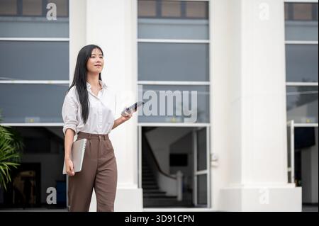 Asian woman in white shirt holding phone and laptop while walking out of university or office building. Student leaving school, Worker get off work. Stock Photo