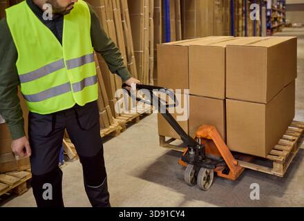 Warehouse worker moving cardboard boxes on pallet jack among storage racks Stock Photo