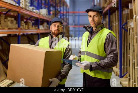 Warehouse staff checking inventory clipboard while coworker carries box in aisle Stock Photo