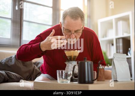 Old white caucasian man wearing glasses smelling coffee aroma from filter paper on the dripper at wooden table. Pour over or Hand Drip. Stock Photo