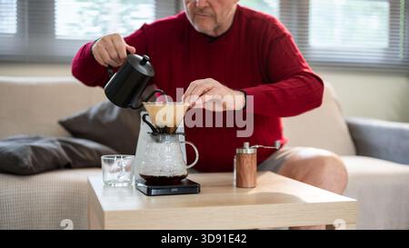 Old white caucasian man using kettle pouring hot water into ground coffee in filter paper on the dripper at wooden table. Pour over or Hand Drip. Stock Photo