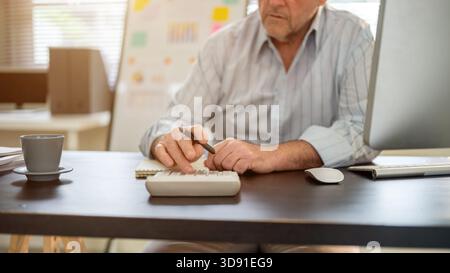 Close up of an old man hands holding a pen and using or pressing on a calculator on working table in his office. Company job, Boss or Senior manager. Stock Photo