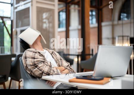 Man in plaid shirt sitting leaning against chair with book covered his face in front of a laptop at cafe table. Study or Working. Sleeping or Resting. Stock Photo