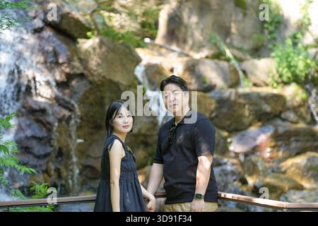 A man and woman are posing for a picture in front of a waterfall. The man is wearing a black shirt and the woman is wearing a black dress. Scene is ro Stock Photo