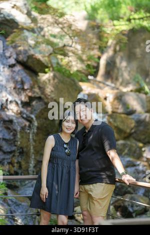 A man and woman are posing for a picture in front of a waterfall. The man is wearing a black shirt and khaki shorts, while the woman is wearing a blac Stock Photo