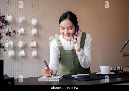 Female barista or owner in green apron talking on phone and holding a pen writing in notebook on counter bar. Cafe or Coffee shop, Small business. Stock Photo