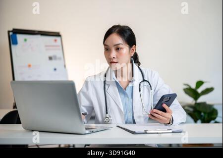 Asian female doctor with stethoscope holding smartphone and looking at laptop on working table in diagnosis room. Hospital or Clinic, Modern office. Stock Photo