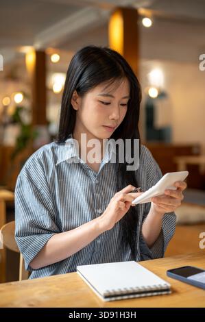 Asian woman is holding and using a calculator with notebook and phone aside sitting at wooden table in the cafe. Income and Expenses. Money management Stock Photo
