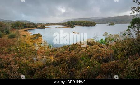 Panoramic view of a remote Kerry lake with autumn bogland, heather, islands, and misty mountains under a cloudy sky. Stock Photo