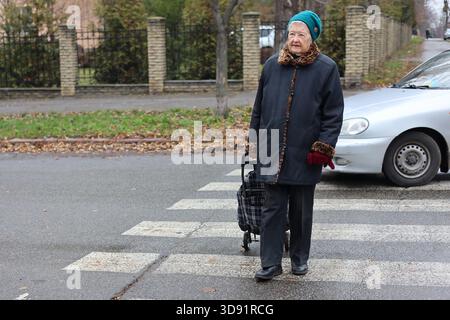 Elderly woman on street. Pensioner walks with cart for things. Woman walks on road. Transportation of personal belongings. Stock Photo