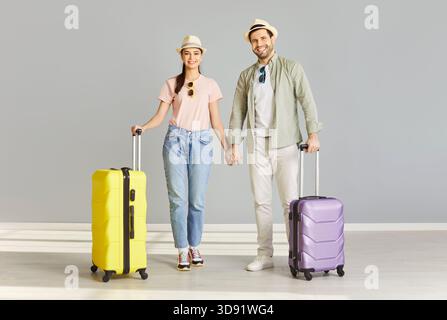 Smiling couple with yellow and purple suitcases holding hands in empty room excited for summer trip. Stock Photo