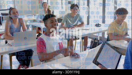 Studying teen male in white-red shirt using laptop and tablet in classroom with binary overlay Stock Photo