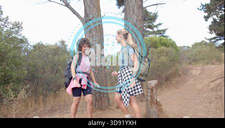 Walking two women talking on dirt trail, carrying backpacks, tied shirts, pine trunks, blue overlay Stock Photo