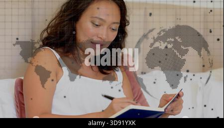 Writing woman in white sleeveless top, holding blue notebook and black pen on bed, globe overlay Stock Photo