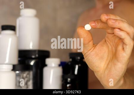 Close-up of hand holding round white tablet with supplement bottles in background. Muscular man holding small white round tablet between fingers, vari Stock Photo
