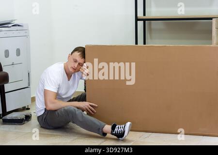 Tired man resting head on arm against cardboard box. Exhausted young man resting his head while sitting against a big cardboard package indoors Stock Photo