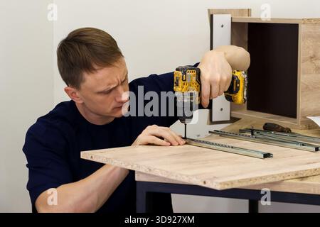 Focused worker drilling into furniture panel with cordless screwdriver. Side view of focused worker using cordless screwdriver to drill into wooden fu Stock Photo