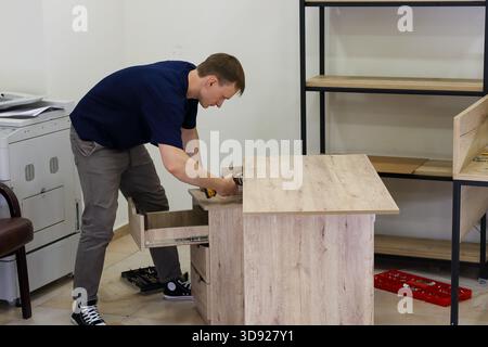Man using drill on desk top during assembly. Leaning man operates cordless drill on wooden desk surface near open drawer, with tools on floor in offic Stock Photo