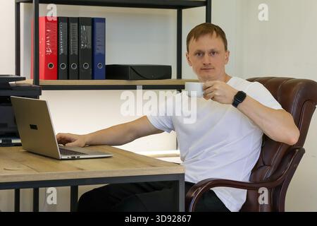 Smiling professional enjoying coffee break, working on laptop at desk in bright office environment with storage. (94 characters) Stock Photo