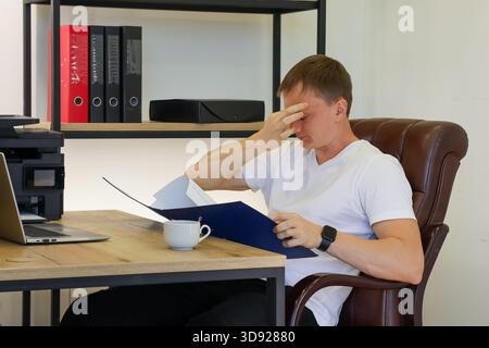 Man in office expresses frustration with hand on forehead while holding open folder of documents at desk with computer and cup. Stock Photo
