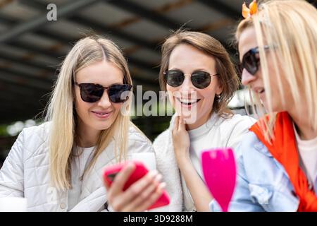 Smiling friends look at phone together, enjoying shared moment with bright expressions and relaxed mood. Casual style, sunglasses and vivid colors Stock Photo
