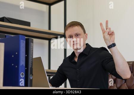 Male professional displaying victory gesture with hand in contemporary office setup with laptop and files Stock Photo