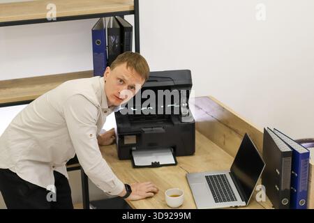 Annoyed office employee stands bent over open printer with hand on desk, expressing irritation in workspace. Stock Photo