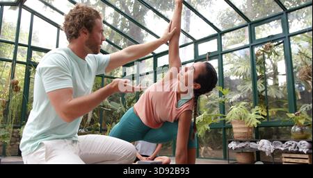 Guiding teacher kneeling beside woman in side plank in greenhouse wearing teal leggings with block Stock Photo