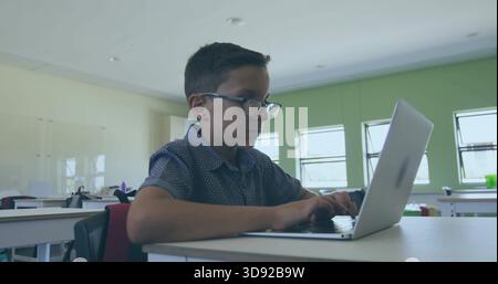 Typing boy wearing glasses and patterned shirt at classroom desk, using open silver laptop Stock Photo