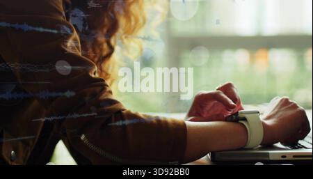 Typing woman wearing brown jacket and light smartwatch, using laptop near window and waveforms Stock Photo
