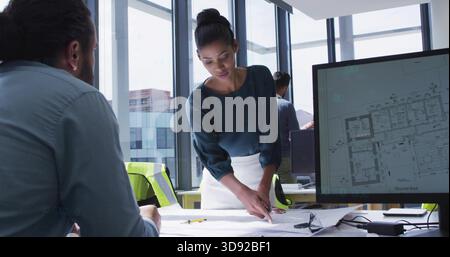 Pointing lead in teal blouse, white skirt leaning over table in office with monitor, vest, plans Stock Photo