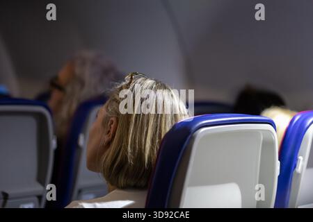 close-up back view of a blonde female passenger with sunglasses resting on her head, seated in an economy class airplane seat in the dark cabin during Stock Photo