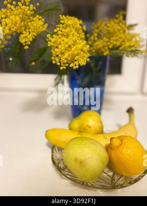 Fresh fruits such as bananas, pears, and lemons are beautifully arranged in a bowl with bright yellow mimosae flowers in a blue vase, enhancing the sc Stock Photo