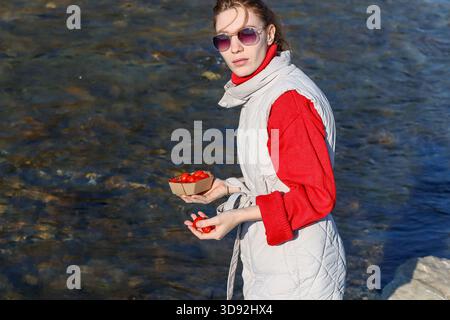A side view of a woman in red sweater and gray vest holding a container of red cherry tomatoes near a flowing river. Stock Photo