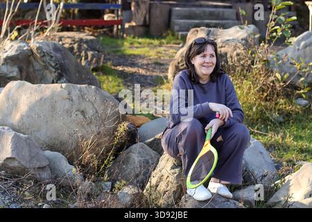 Mature woman in blue outfit sits on large stones, smiling while holding yellow and green badminton racket in grassy area with path. Stock Photo