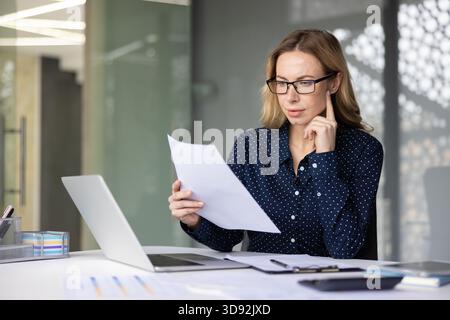 Young businesswoman wearing glasses focusing on financial documents while sitting at her desk with a laptop, clipboard, and pens in a contemporary office environment Stock Photo