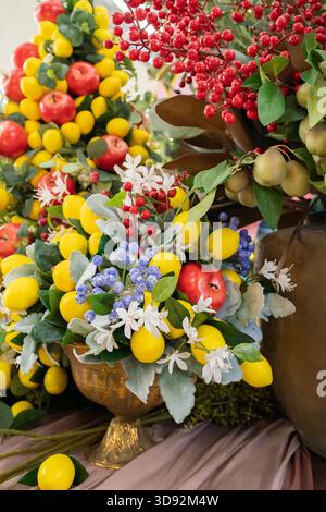 A lush centerpiece featuring bright yellow lemons, red berries, purple flowers, and green foliage arranged in an ornate gold pedestal vase. Stock Photo