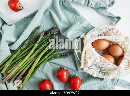 A bunch of fresh asparagus lies next to a reusable mesh bag filled with brown eggs, accented by ripe cherry tomatoes on a light-colored surface with a blue cloth. The setting suggests preparation for a healthy, home-cooked meal. Stock Photo