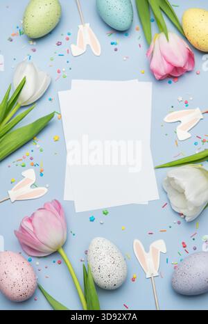 Easter greeting card surrounded by colorful eggs, tulips and bunny ears on pastel blue background with sprinkles Stock Photo