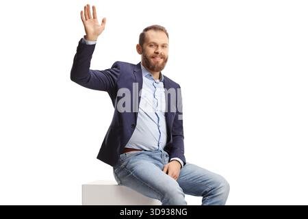 Man in suit and jeans sitting on a white cube and waving isolated on white background Stock Photo