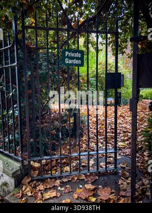 A black metal gate with a 'Private Garden' sign opening onto a path covered in autumn leaves.  London. UK Stock Photo