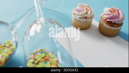 Showing two frosted cupcakes on card at pale blue surface, glasses spilling sprinkles, copy space Stock Photo