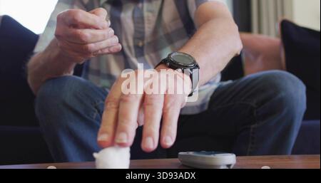 Handling lancet, senior male in plaid shirt, on sofa reaching cotton ball on table with glucometer Stock Photo