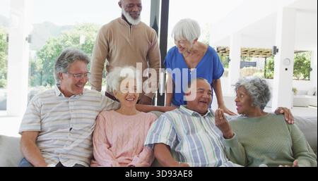 Smiling senior group chatting on light-colored sofa near glass sliding doors, wearing sweaters Stock Photo