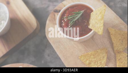Showing white bowl of chunky tomato salsa with rosemary on wooden board, dark slate, nachos, salt Stock Photo
