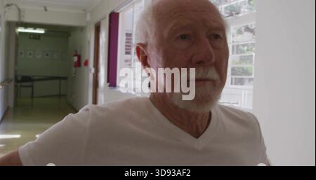 Standing senior man in white V-neck tee gazing down care home corridor with windows, extinguisher Stock Photo